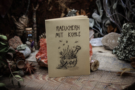 Kleines geschlossenes Zine "Raeuchern mit Kohle"" , Anleitung mit Bildern zum Räuchern, das Papier sieht gelblich aus mit schwarzer Schrift.aufgestellt auf einem dunklen Tisch umgeben von Kräutern und Kristallen