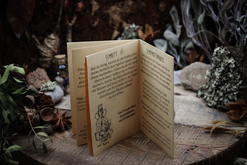 Kleines Zine, Anleitung mit Bildern zum Räuchern, das Papier sieht gelblich aus mit schwarzer Schrift.aufgestellt auf einem dunklen Tisch umgeben von Kräutern und Kristallen