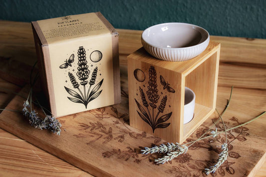 Eine Duftlampe aus Holz und grau-beiger Keramik. In das Holz eingebrannt ist das Motiv eines Lavendels, daneben der Mond, eine Motte und kleine Sterne. Neben der Lampe steht der Umkarton auf den das selbe Motiv gedruckt ist.