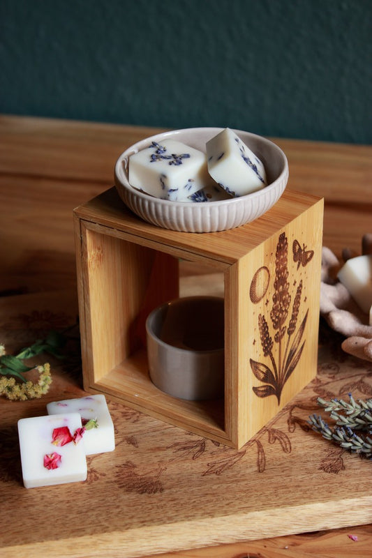 Eine Duftlampe aus Holz und grau-beiger Keramik. In das Holz eingebrannt ist das Motiv eines Lavendels, daneben der Mond, eine Motte und kleine Sterne. In der Keramikschale liegt Duftwachs mit Lavendelblüten.