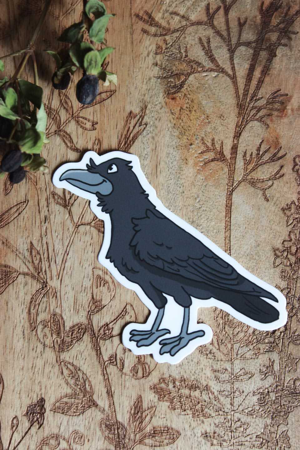 Hochkant: Das Bild zeigt einen Aufkleber auf einer Holzoberfläche mit kunstvollen Pflanzenmustern. Der Aufkleber selbst stellt einen schwarzen Vogel dar, einen Raben, der detailliert gestaltet ist, mit einem auffälligen Schnabel, Federn und Füßen. Der Vogel hat eine weiße Umrandung, die ihn hervorhebt. In der oberen linken Ecke des Bildes sind außerdem getrocknete dunkle Beeren zu sehen.
