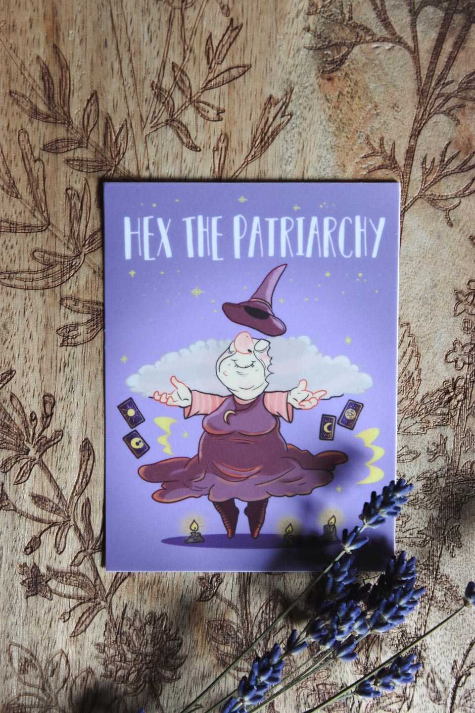Das hochgeladene Bild zeigt eine Cartoon-Illustration einer älteren Frau, die als Hexe gekleidet in der Luft schwebt und einen zufriedenen Gesichtsausdruck hat. Sie trägt ein lilafarbenes Kleid sowie einen spitzen Hut, der über ihrem Kopf schwebt. Um sie herum sind vier Tarotkarten zu sehen sowie drei brennende Kerzen, die auf dem Boden stehen. Der Hintergrund ist in einem hellen Lilaton gehalten und zeigt Sterne und Wolken.
Oben im Bild steht in großen weißen Großbuchstaben „HEX THE PATRIARCHY“.