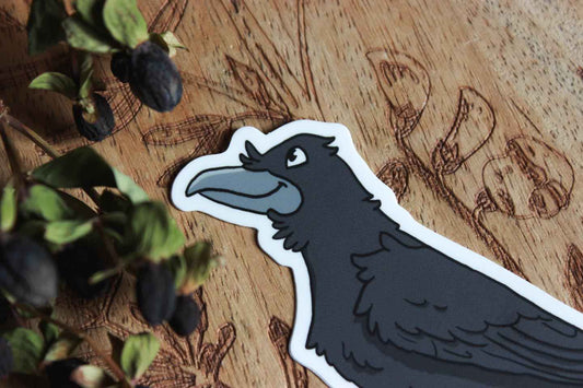 Nahaufnahme: Das Bild zeigt einen Aufkleber auf einer Holzoberfläche mit kunstvollen Pflanzenmustern. Der Aufkleber selbst stellt einen schwarzen Vogel dar, einen Raben, der detailliert gestaltet ist, mit einem auffälligen Schnabel, Federn und Füßen. Der Vogel hat eine weiße Umrandung, die ihn hervorhebt. In der oberen linken Ecke des Bildes sind außerdem getrocknete dunkle Beeren zu sehen.
