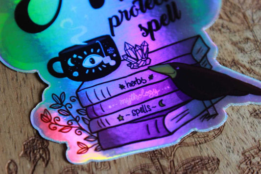 Nahaufnahme: Das Bild zeigt einen Aufkleber, der auf einer Holzoberfläche mit floralen Gravuren liegt. Der Aufkleber ist holografisch. Darauf steht der Text: „No is a protection spell.“ Unter dem Text sind Illustrationen zu sehen, darunter eine Tasse mit einem Augendesign, drei übereinander gestapelte Bücher mit den Titeln „herbs“ „mythology“ und „spells“ sowie ein schwarzer Vogel, der neben den Büchern sitzt. Zudem gibt es kleine Pflanzen und Kristalle um die Bücher herum.