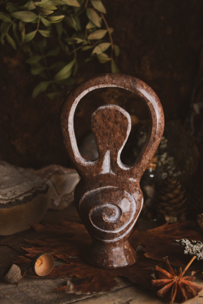 Eine kunstvoll gestaltete Skulptur, Göttinenfigur, dunkelbraun und poliert präsentiert in einer natürlichen Umgebung mit getrockneten Blättern und Sternanis.