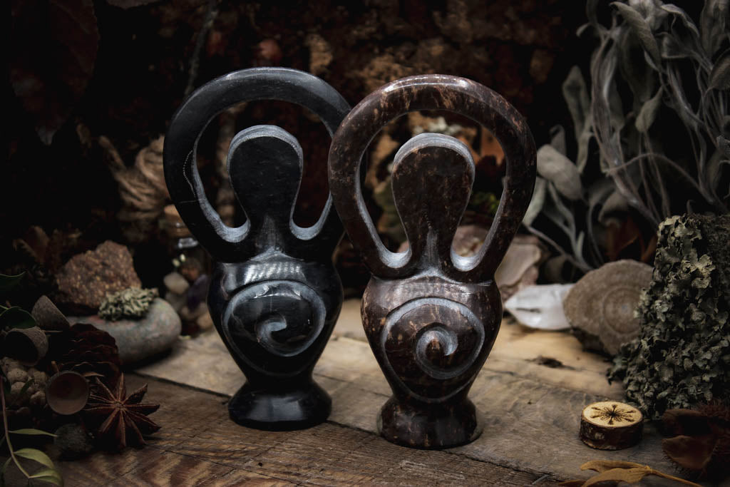 Zwei kunstvoll gestaltete Skulpturen, Göttinenfigur, dunkel und poliert präsentiert in einer natürlichen Umgebung mit getrockneten Blättern und Sternanis.