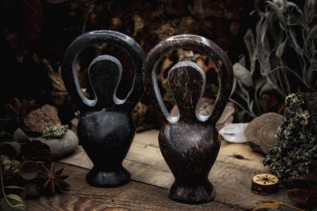 Zwei kunstvoll gestaltete Skulpturen, Göttinenfigur, dunkelbraun und poliert präsentiert in einer natürlichen Umgebung mit getrockneten Blättern und Sternanis.