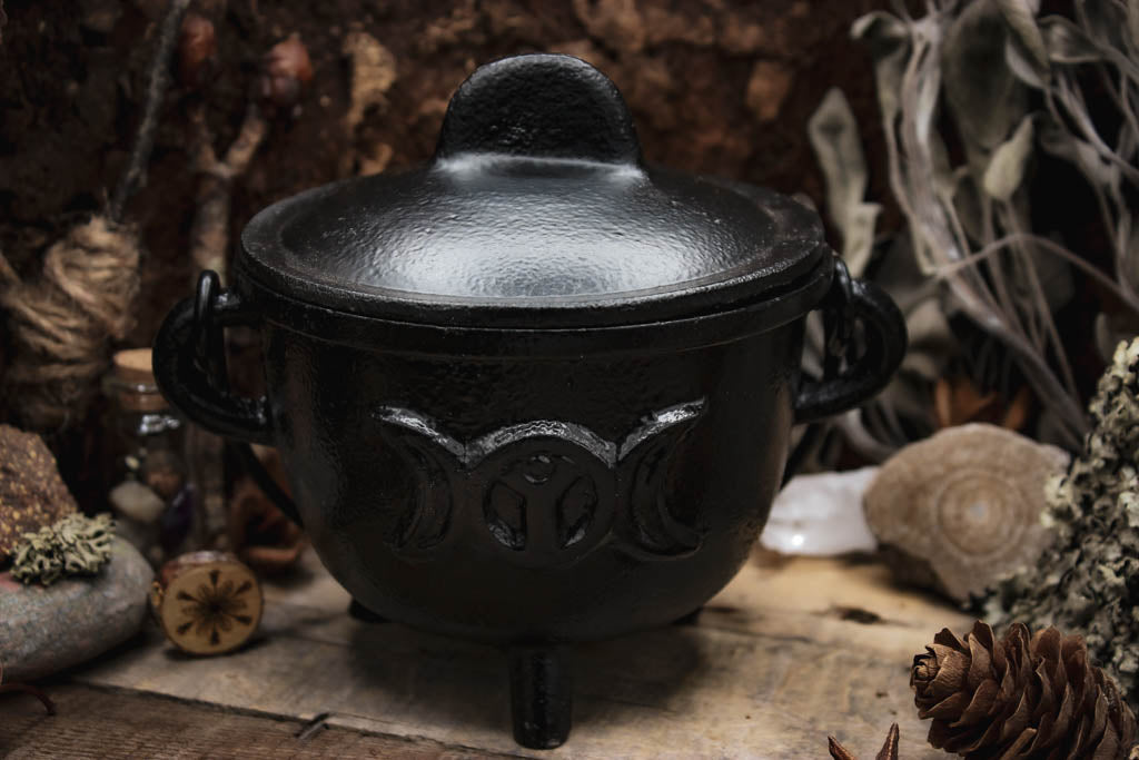 Rustikaler schwarzer Gusseisen-Topf geschlossen mit einem passenden Deckel, umgeben von natürlichen Elementen wie Kiefernzapfen, getrockneten Blumen und Gewürzen auf einem hölzernen Hintergrund, Auf dem Kessel sieht man den Mond in verschiedenen Phasen