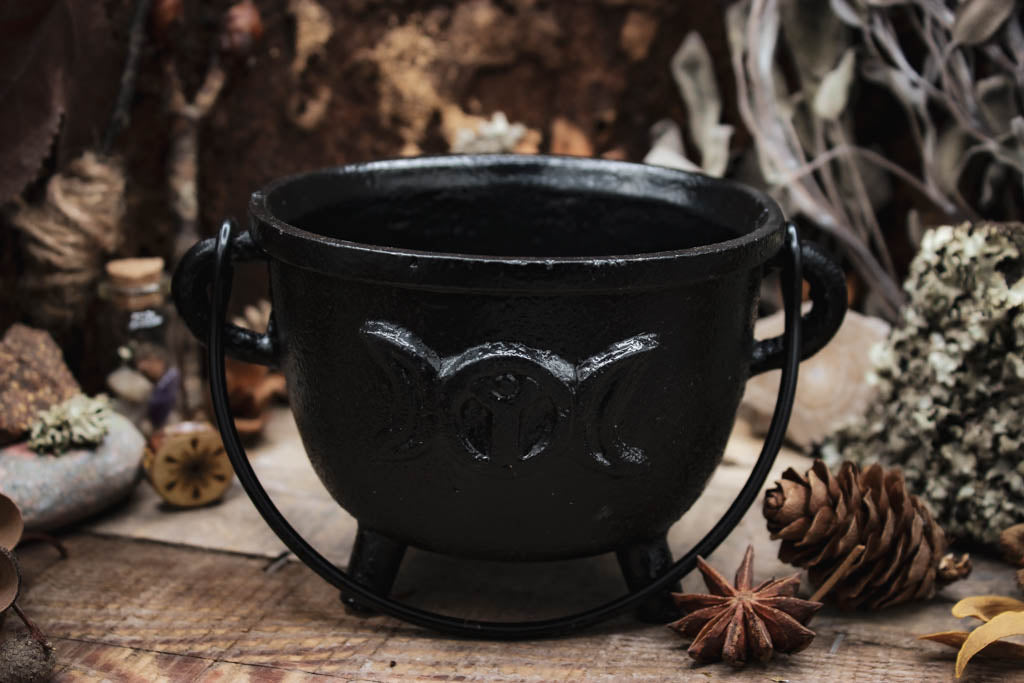 Rustikaler schwarzer Gusseisen-Topf leer, umgeben von natürlichen Elementen wie Kiefernzapfen, getrockneten Blumen und Gewürzen auf einem hölzernen Hintergrund, Auf dem Kessel sieht man den Mond in verschiedenen Phasen