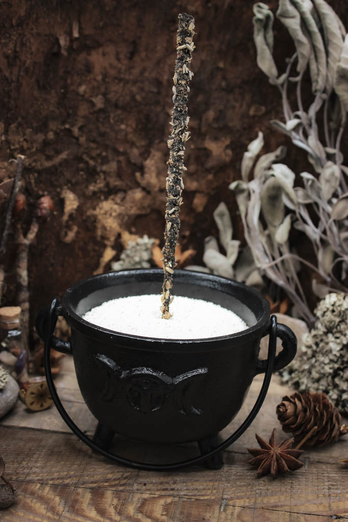 Rustikaler schwarzer Gusseisen-Topf gefüllt mit weißem Sand, ein Räucherstick steht darin, umgeben von natürlichen Elementen wie Kiefernzapfen, getrockneten Blumen und Gewürzen auf einem hölzernen Hintergrund, Auf dem Kessel sieht man den Mond in verschiedenen Phasen