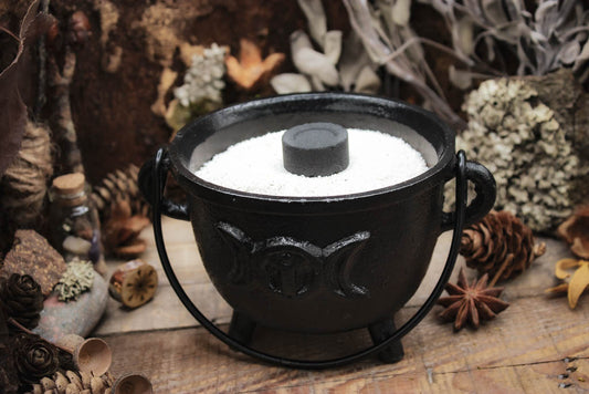 Rustikaler schwarzer Gusseisen-Topf gefüllt mit weißem Sand, umgeben von natürlichen Elementen wie Kiefernzapfen, getrockneten Blumen und Gewürzen auf einem hölzernen Hintergrund, Auf dem Kessel sieht man den Mond in verschiedenen Phasen