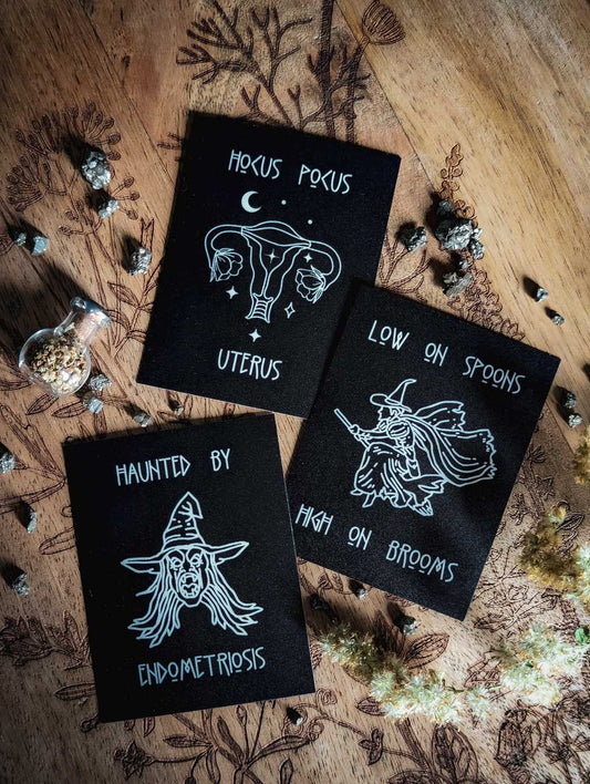Auf einem Holzbrett liegen drei mattschwarze Sticker. Auf dem oberen Sticker ist in weißer Farbe ein Uterus mit Blüten, Sternen und Mond zu sehen. Dazu der Spruch: Hocus Pocus Uterus. Der zweite Sticker zeigt eine Hexe auf einem Besen. Dazu der Spruch: Low on Spoons High in Brooms. Der dritte Sticker zeigt eine Fratze mit aufgerissenen Mund, wirren langen Haaren und Hexenhut. Dazu der Spruch: Haunted by endometriosis.