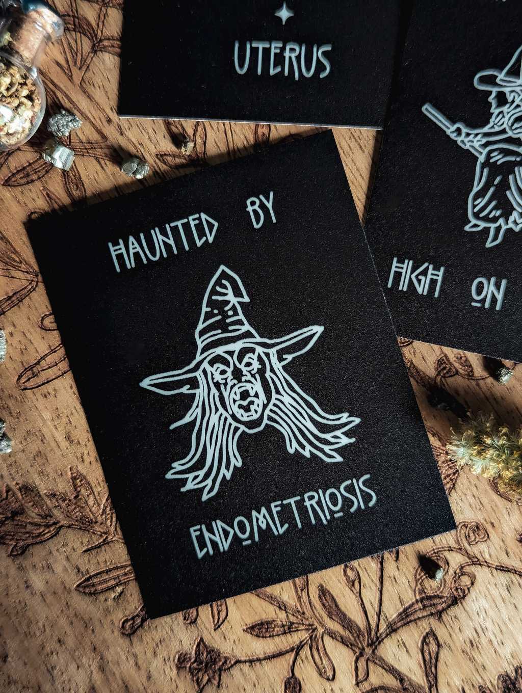 Auf einem Holzbrett liegt ein mattschwarzer Sticker. In weißer Farbe ist eine Fratze mit aufgerissenen Mund, wirren langen Haaren und Hexenhut zu sehen. Dazu der Spruch: Haunted by endometriosis.