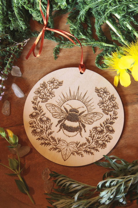 Auf einem Holztisch liegen verschiedene Kräuter wie Schafgarbe, Wermut und Johanniskraut um ein Wandbild aus Holz drumherum. Das Holz-Ornament zeigt eine Hummel. Eingerahmt wird sie von Johanniskraut, Kamille und einem Efeublatt.
