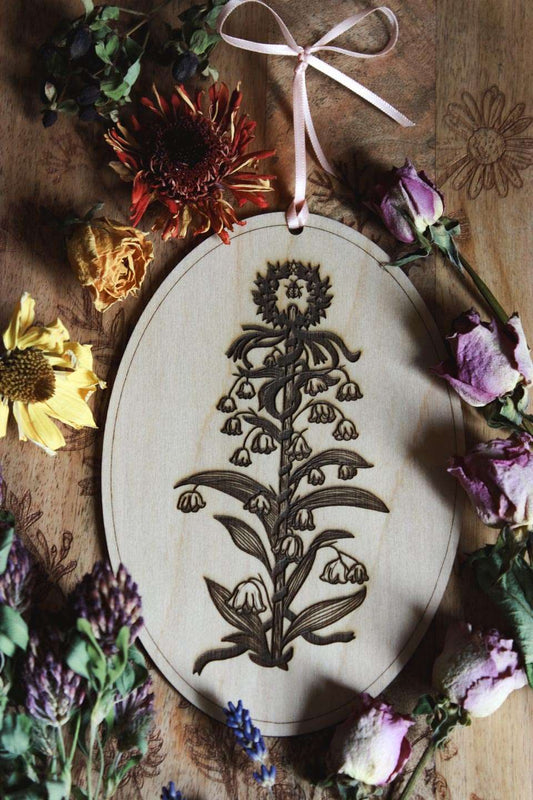 Das Bild zeigt eine ovale Holzplatte mit einem eingravierten floralen Design, umgeben von verschiedenen getrockneten Blumen. Das Holzstück hat oben ein rosafarbenes Band, das zum Aufhängen dient. Das eingravierte Design zeigt einen Maibaum, der von Maiglöckchen umschlungen wird. Oben an der Spitze ist ein winziges Pentagramm und ein Marienkäfer zu sehen.