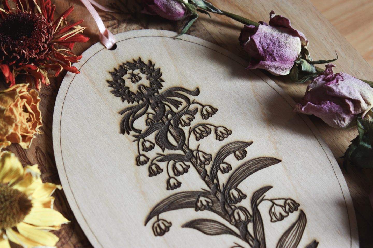 Das Bild zeigt eine ovale Holzplatte mit einem eingravierten floralen Design, umgeben von verschiedenen getrockneten Blumen. Das Holzstück hat oben ein rosafarbenes Band, das zum Aufhängen dient. Das eingravierte Design zeigt einen Maibaum, der von Maiglöckchen umschlungen wird. Oben an der Spitze ist ein winziges Pentagramm und ein Marienkäfer zu sehen.