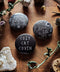 Drei runde Buttons mit mystischen Katzenmotiven, arrangiert auf einer hölzernen Fläche zwischen getrockneten Blüten, Kristallen und kleinen Naturfundstücken. Ein Button zeigt eine schwarze Katze mit Pentagramm, einer trägt eine Hexenhut‑Katze im Mondschein, und der dritte zeigt den Schriftzug ‚Cozy Cat Coven‘.