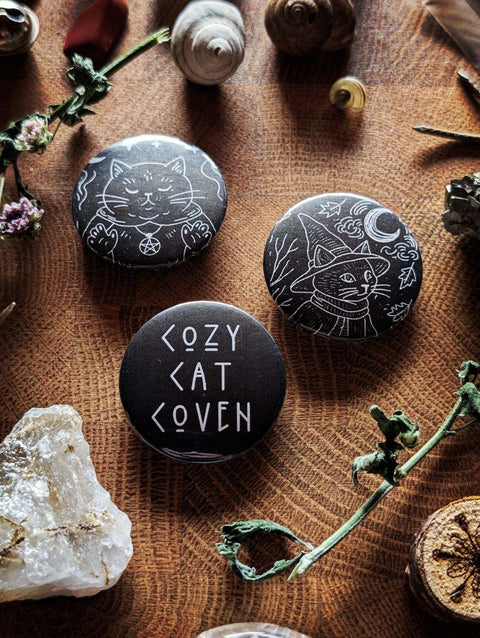 Drei runde Buttons mit mystischen Katzenmotiven, arrangiert auf einer hölzernen Fläche zwischen getrockneten Blüten, Kristallen und kleinen Naturfundstücken. Ein Button zeigt eine schwarze Katze mit Pentagramm, einer trägt eine Hexenhut‑Katze im Mondschein, und der dritte zeigt den Schriftzug ‚Cozy Cat Coven‘.
