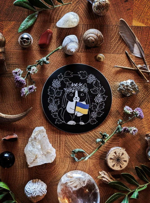 Ein runder schwarzer Sticker mit Motiv. Das Motiv zeigt eine schwarz-weiße niedliche Katze mit Blumenkrone auf dem Kopf. Sie hält die blau-gelbe Flagge der Ukraine in der Pfote. Umrahmt wird sie vom Schneeball-Strauch.
