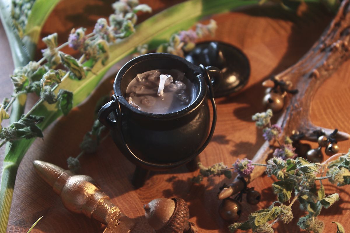 Handgegossene Ritualkerze in einem schwarzen Mini-Kessel, verziert mit Bergkristallstücken. Die Kerze steht auf einer rustikalen Holzoberfläche, umgeben von dekorativen Elementen wie getrockneten Blüten und grünen Blättern. Ideal für spirituelle Rituale, Meditation oder als magisches Wohnaccessoire.