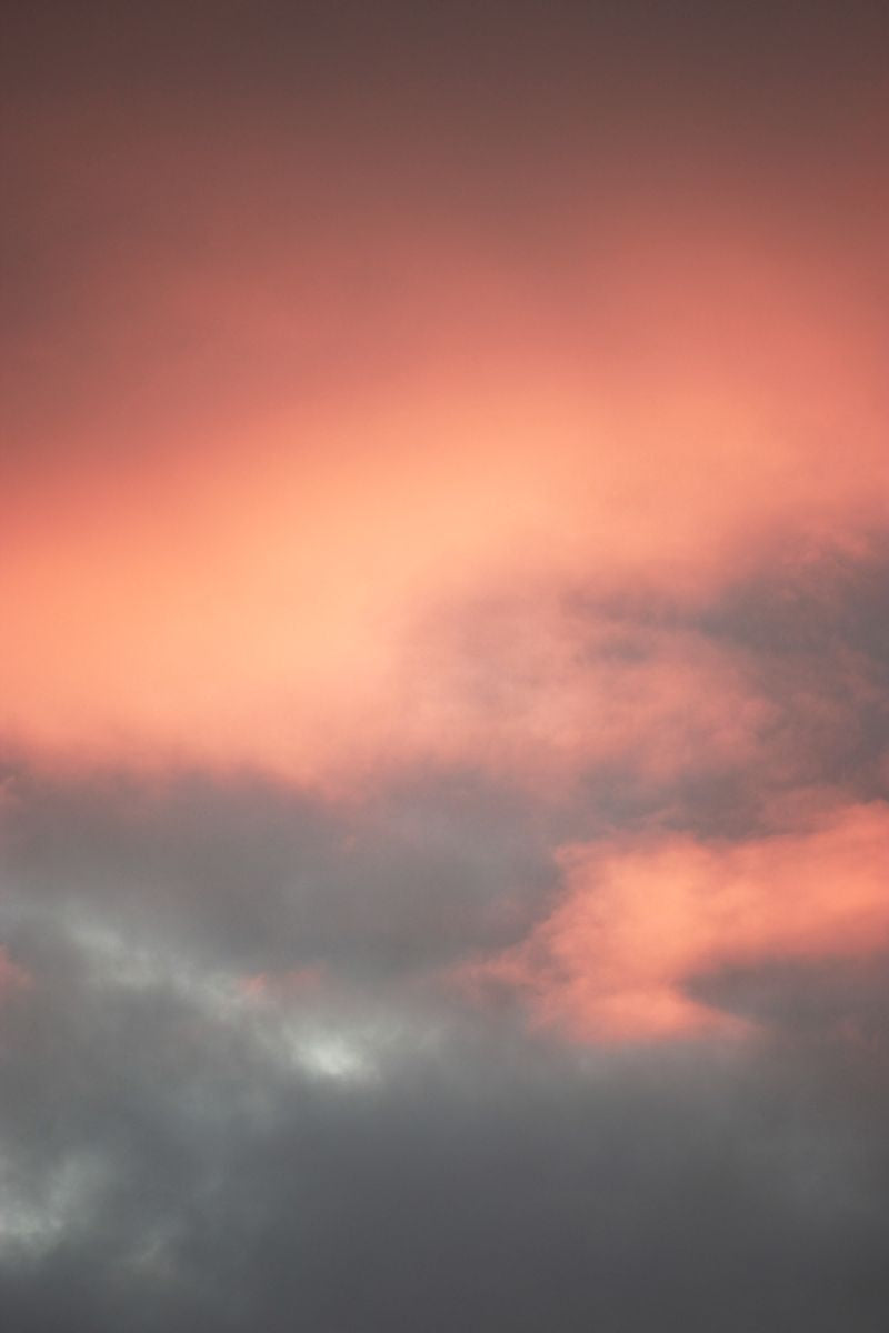 Stimmungsvolles Himmelsbild mit wolkenverhangenem Sonnenlicht in warmen Rosa- und Orangetönen, das an die Farbverwandlung der Ritualkerze erinnert – von grauer Ruhe im unangezündeten Zustand bis hin zu rötlicher Wärme beim Brennen.