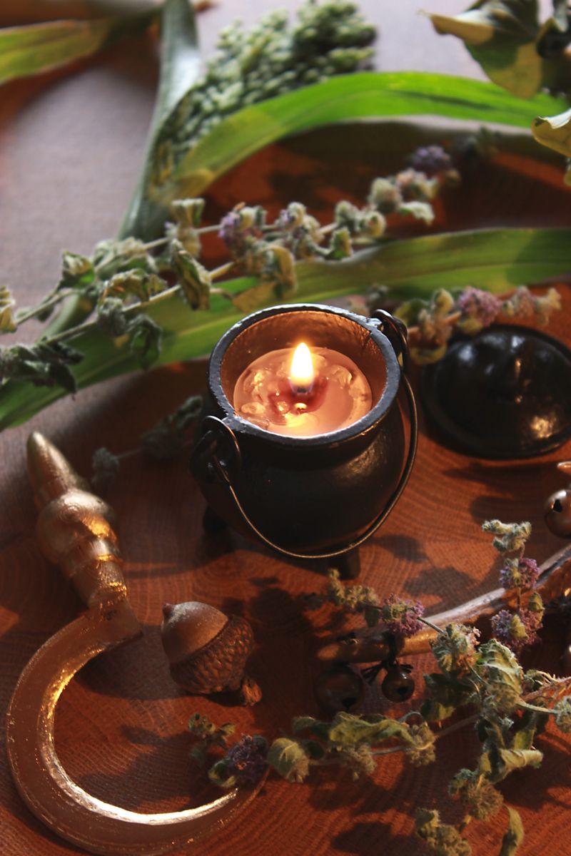 Handgegossene Ritualkerze in einem schwarzen Mini-Kessel, verziert mit Bergkristallstücken. Sie brennt und taucht die Umgebung in warmes Licht. Die Kerze steht auf einer rustikalen Holzoberfläche, umgeben von dekorativen Elementen wie getrockneten Blüten, einer Sichel und grünen Blättern. Ideal für spirituelle Rituale, Meditation oder als magisches Wohnaccessoire.