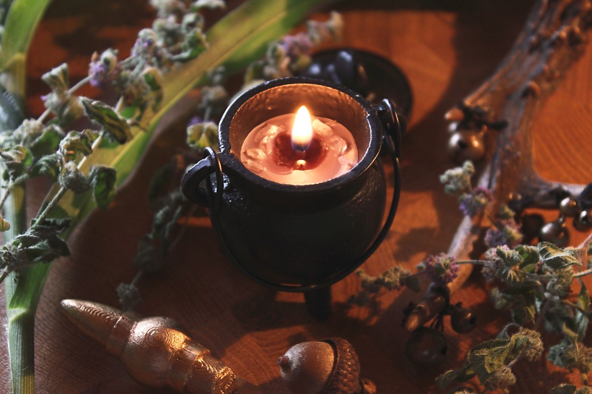 Handgegossene Ritualkerze in einem schwarzen Mini-Kessel, verziert mit Bergkristallstücken. Sie brennt und taucht die Umgebung in warmes Licht. Die Kerze steht auf einer rustikalen Holzoberfläche, umgeben von dekorativen Elementen wie getrockneten Blüten und grünen Blättern. Ideal für spirituelle Rituale, Meditation oder als magisches Wohnaccessoire.