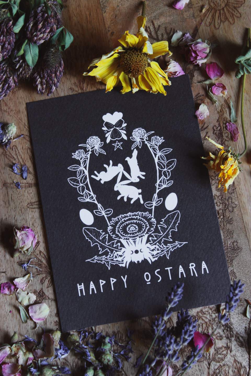 Das Bild zeigt eine schwarze Karte mit weißen Illustrationen und dem Text „HAPPY OSTARA“ unten. Um die Karte herum liegen verschiedene getrocknete Blumen und Kräuter, darunter Klee, Lavendel und Rosen. Auf der Karte sind drei Hasen, Eier und florale Elemente in einem symmetrischen Design abgebildet. Die Karte liegt auf einer Holzoberfläche mit weiteren floralen Gravuren.
