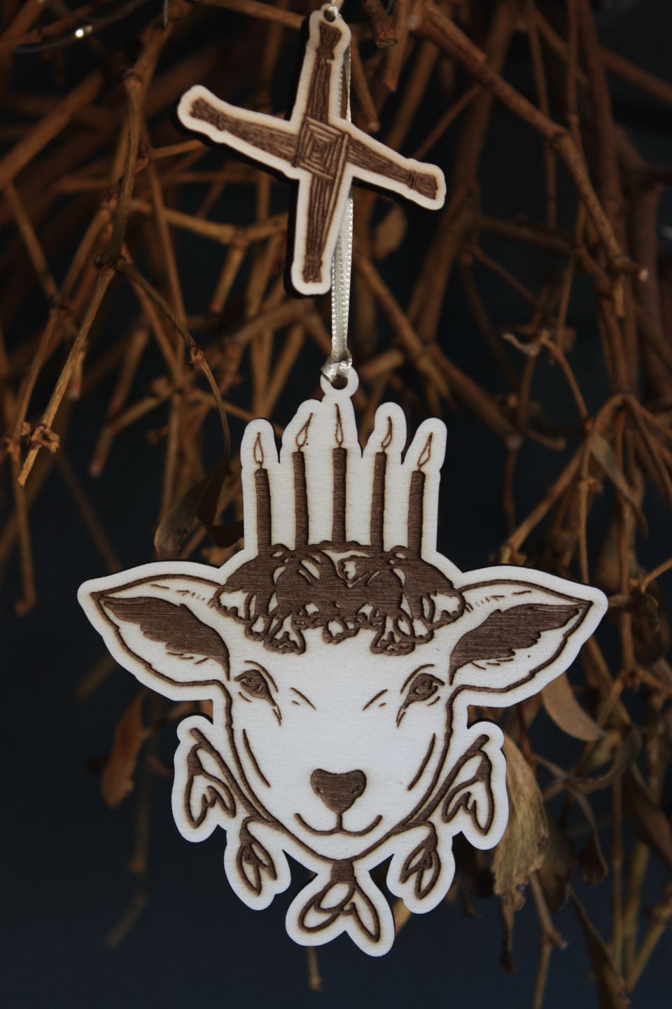 Nahaufnahme: An einem alten Mistelzweig vor einer dunklen blau-grünen Wand hängt an einem hellgelben Band ein Ornament aus Holz. Es zeigt den Kopf eines Schafs mit fünf Kerzen auf dem Haupt, die auf Schlüsselblumen stehen. Unter dem Kopf befinden sich fünf Blüten des Schneeglöckchens. Als kleineres weiteres Holz-Ornament hängt über dem Schaf das Kreuz der Brigid.