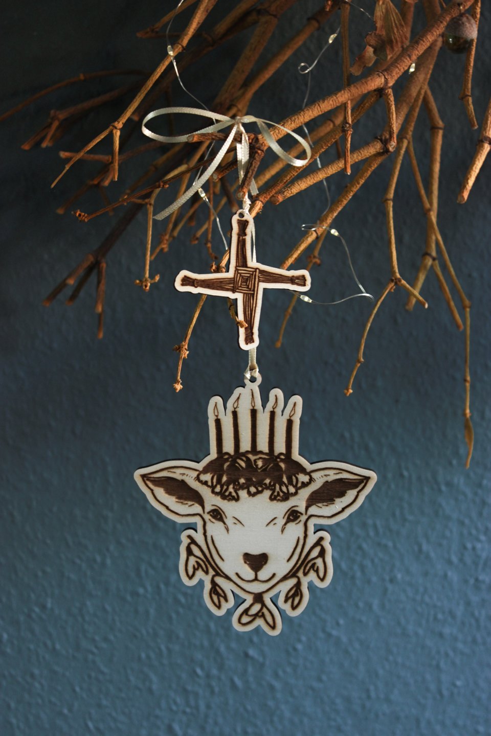Hochkant: An einem alten Mistelzweig vor einer dunklen blau-grünen Wand hängt an einem hellgelben Band ein Ornament aus Holz. Es zeigt den Kopf eines Schafs mit fünf Kerzen auf dem Haupt, die auf Schlüsselblumen stehen. Unter dem Kopf befinden sich fünf Blüten des Schneeglöckchens. Als kleineres weiteres Holz-Ornament hängt über dem Schaf das Kreuz der Brigid.