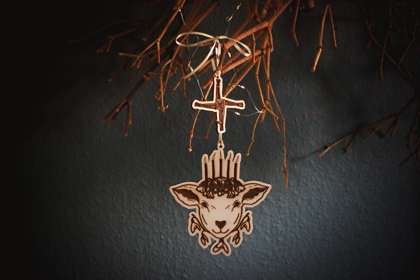 An einem alten Mistelzweig vor einer dunklen blau-grünen Wand hängt an einem hellgelben Band ein Ornament aus Holz. Es zeigt den Kopf eines Schafs mit fünf Kerzen auf dem Haupt, die auf Schlüsselblumen stehen. Unter dem Kopf befinden sich fünf Blüten des Schneeglöckchens. Als kleineres weiteres Holz-Ornament hängt über dem Schaf das Kreuz der Brigid.