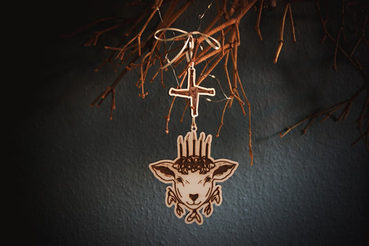 An einem alten Mistelzweig vor einer dunklen blau-grünen Wand hängt an einem hellgelben Band ein Ornament aus Holz. Es zeigt den Kopf eines Schafs mit fünf Kerzen auf dem Haupt, die auf Schlüsselblumen stehen. Unter dem Kopf befinden sich fünf Blüten des Schneeglöckchens. Als kleineres weiteres Holz-Ornament hängt über dem Schaf das Kreuz der Brigid.
