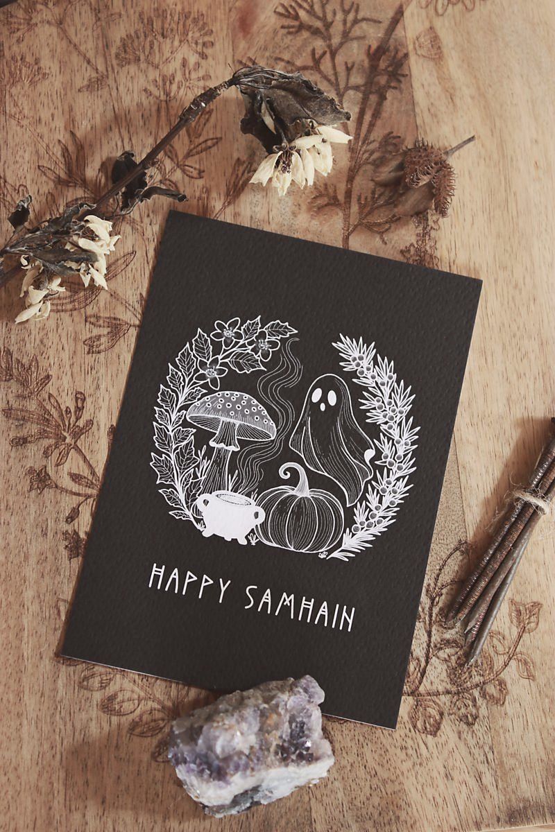 Eine schwarze Postkarte mit weißem Druck. Es zeigt folgendes Motiv: Neben Geist, Kürbis und Fliegenpilz steht ein Hexenkessel aus dem Rauch empor steigt. Eingerahmt ist das Motiv von schwarzem Bilsenkraut und Wacholder, die typisch für die Samhain-Nacht sind. Darunter steht: HAPPY SAMHAIN