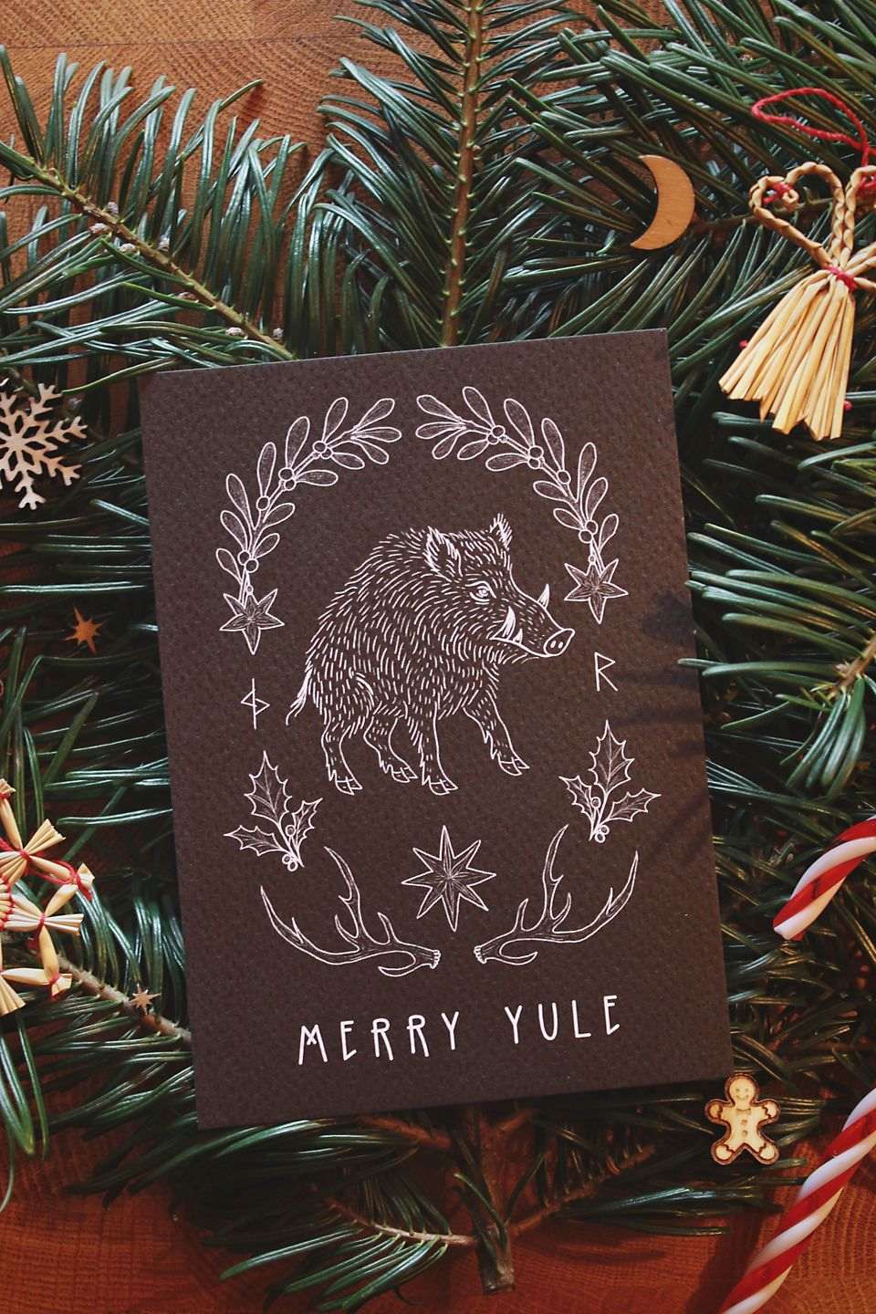 Hochkant: Gebettet auf Tannenzweigen und weihnachtliche Dekoration liegt in der Mitte eine schwarze Karte mit weißem Aufdruck. Diese Grußkarte zu Jul (Wintersonnenwende) zeigt ein Wildschwein. Eingerahmt wird es von typischen Jul-Motiven wie Mistelzweigen, Stechpalmen, Strohsternen, Geweihen und passenden Runen. Darunter steht: Merry Yule