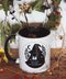 Die Tasse aus Keramik trägt das Motiv "Stay Safe Be Kind Hex The Patriarchy". Die vermummte Hexen-Gestalt mit Hexen-Hut, hält eine Katze in den Armen. Auf ihrer Schulter ist ein kleines Pentagramm zu sehen. Um sie herum sind Salbei und Beifuß. Das Motiv ist in schwarz gehalten, genau so wie der Henkel und das Innere der Tasse. Ansonsten ist die Tasse weiß. Umrahmt wird die Tasse von vielen Pflanzen.