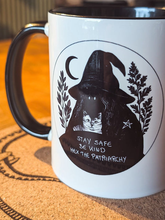 Nahaufnahme der Tasse:
Die Tasse aus Keramik trägt das Motiv "Stay Safe Be Kind Hex The Patriarchy". Die vermummte Hexen-Gestalt mit Hexen-Hut, hält eine Katze in den Armen. Auf ihrer Schulter ist ein kleines Pentagramm zu sehen. Um sie herum sind Salbei und Beifuß. Das Motiv ist in schwarz gehalten, genau so wie der Henkel und das Innere der Tasse. Ansonsten ist die Tasse weiß.