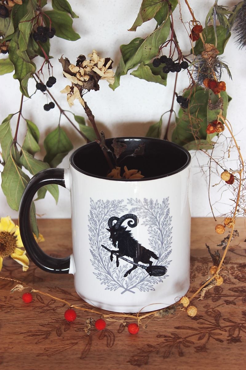 Umrahmt von Pflanzen steht eine weiße Tasse mit schwarzem Inneren und Henkel auf einem Holzbrett. Auf ihr zuu sehen ist das Motiv eines schwarzen Ziegenbocks, der auf einem Hexenbesen reitet. Umrahmt ist er von Beifuß.