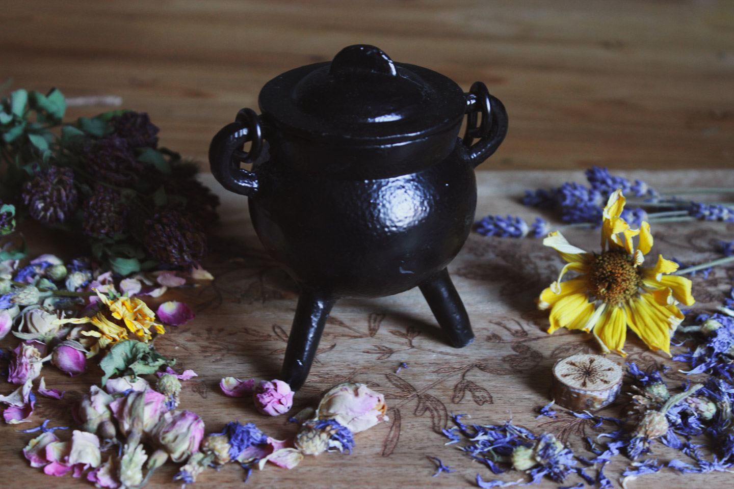 Das Bild zeigt einen kleinen schwarzen Kessel mit Deckel, der auf drei Beinen steht. Der Kessel ist umgeben von verschiedenen getrockneten Blumen und Kräutern, darunter gelbe, violette und pinkfarbene Blumen.