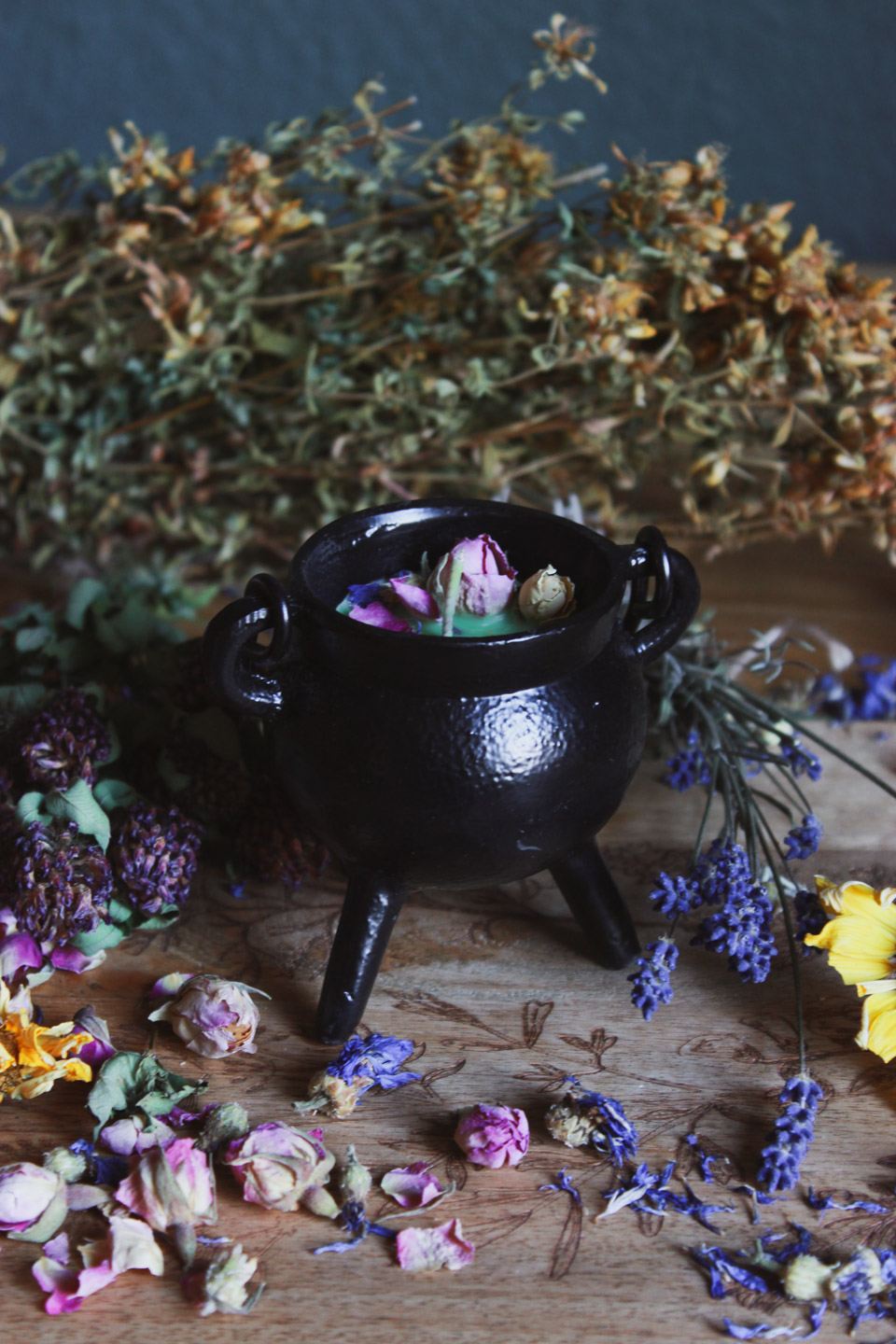 Das Bild zeigt einen kleinen schwarzen Kessel, der auf einer Holzoberfläche steht. Umgeben ist der Kessel von verschiedenen getrockneten Blumen und Kräutern, darunter rosafarbene Rosenknospen, gelbe und lila Blüten. Innerhalb des Kessels befinden sich einige getrocknete Rosenknospen. Im Hintergrund sind weitere getrocknete Pflanzen und Kräuter verstreut, was eine rustikale und natürliche Atmosphäre schafft.