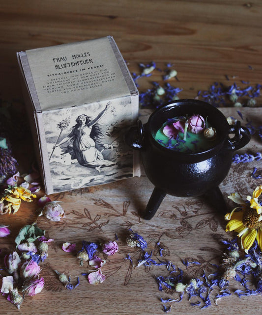 Das Bild zeigt einen kleinen schwarzen Kessel, der mit einer grünen Kerze gefüllt ist. Die Oberfläche ist liebevoll mit getrockneten Blumen dekoriert. Neben dem Kessel liegt eine Schachtel mit der Aufschrift „Frau Holles Blütenfeuer“.