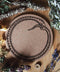 Das Bild zeigt eine Korkunterlage mit dem schwarzen Symbol einer sich selbst verschlingenden Schlange, bekannt als Ouroboros. Sie liegt auf einer Holzfläche und ist von verschiedenen getrockneten Kräutern und Blumen umgeben, darunter Lavendel, Salbei und weitere botanische Elemente.