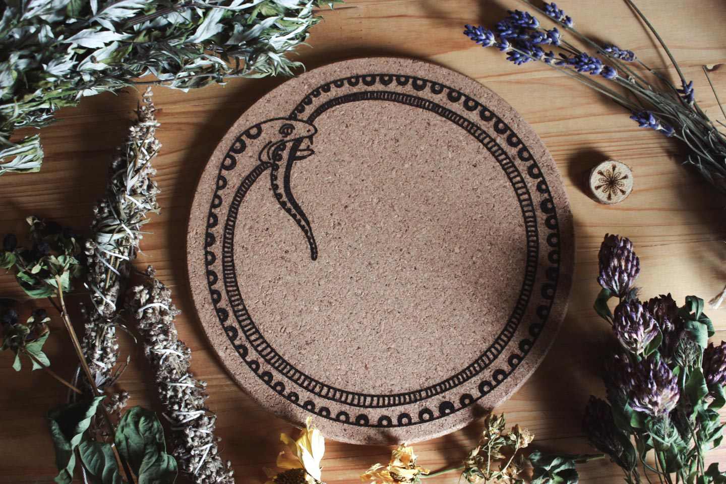 Das Bild zeigt eine Korkunterlage mit dem schwarzen Symbol einer sich selbst verschlingenden Schlange, bekannt als Ouroboros. Sie liegt auf einer Holzfläche und ist von verschiedenen getrockneten Kräutern und Blumen umgeben, darunter Lavendel, Salbei und weitere botanische Elemente.
