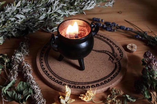 Das Bild zeigt eine Korkunterlage mit dem schwarzen Symbol einer sich selbst verschlingenden Schlange, bekannt als Ouroboros. Sie liegt auf einer Holzfläche und ist von verschiedenen getrockneten Kräutern und Blumen umgeben, darunter Lavendel, Salbei und weitere botanische Elemente. Auf dem Kork steht ein kleiner Kessel mit brennender Kerze darin.