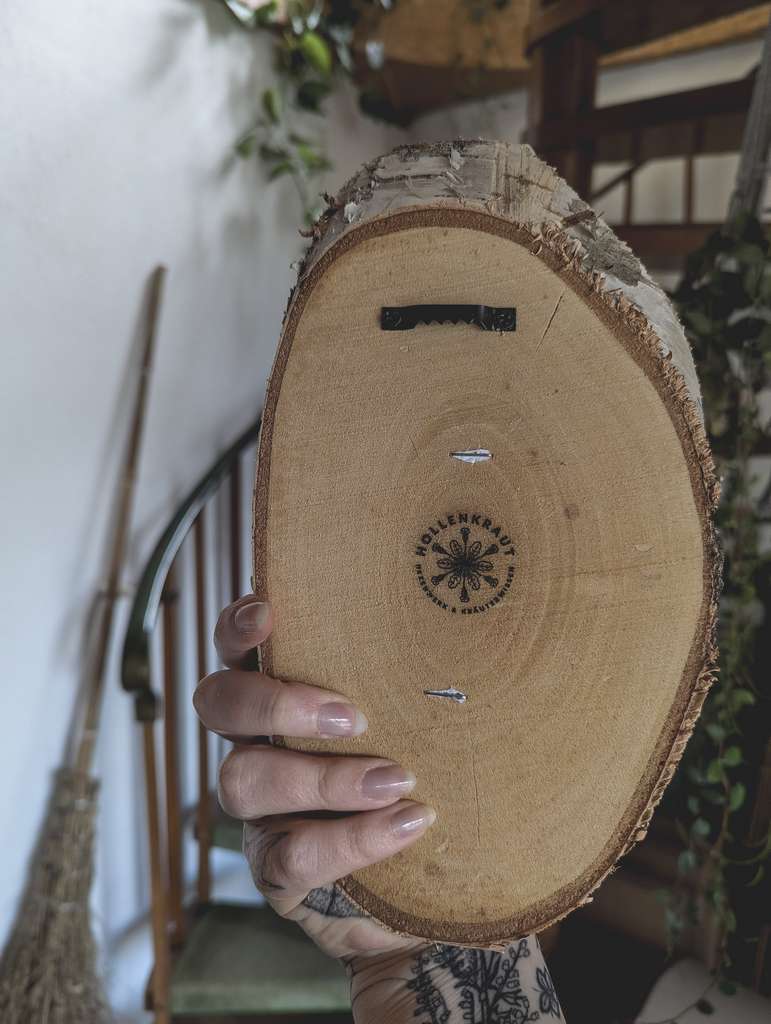 Eine Hand hält eine ovale Holzscheibe in die Kamera. Es ist die Rückseite des Produkts zu sehen. Darauf ist eine Bildaufhängung geschraubt und das Hollenkraut-Logo ist eingebrannt.