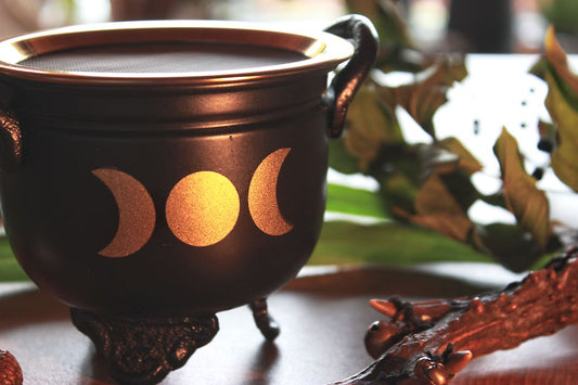 Nahaufnahme: Schwarzes Räuchergefäß in Kessel-Form mit goldenen Akzenten und Triple-Mond-Symbol. Mit Siebeinsatz, dekorativen Griffen und drei verzierten Standfüßen. Umgeben von natürlichen Elementen wie Blättern, Beerenzweig und getrockneten Blüten – ideal für spirituelle Rituale und stilvolle Dekoration.