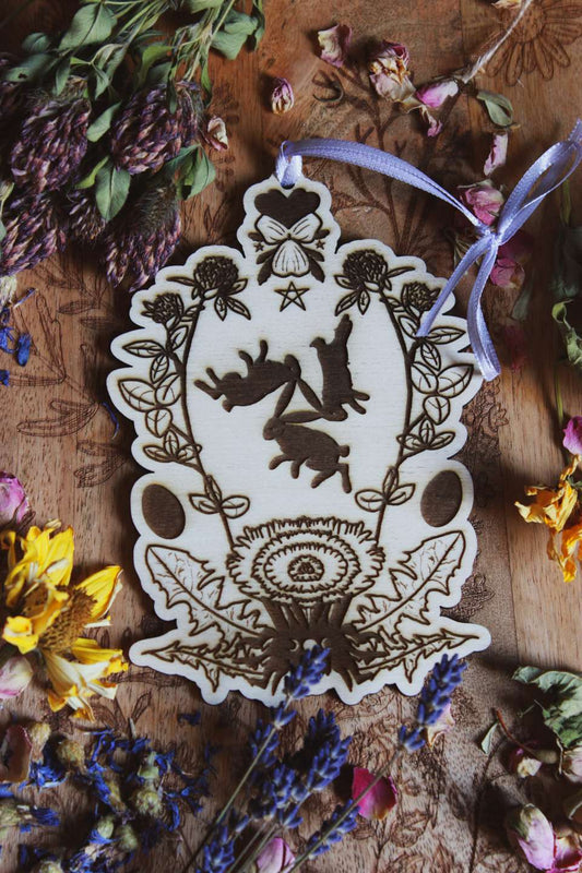 Das Bild zeigt ein kunstvolles Holzornament mit detailliertem Motiv und einer fliederfarbenen Schleife, die oben gebunden ist. Im Zentrum des Ornaments sind drei Hasen dargestellt, die im Kreis laufen, umgeben von floralen Mustern, wie Rotklee, Löwenzahn und Veilchen. Das Ornament liegt auf einer Holzoberfläche, umgeben von verschiedenen getrockneten Blumen, wie Kleeblüten, gelben Blumen und Lavendel.