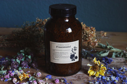 Das Bild zeigt ein dunkelbraunes Glas mit schwarzem Deckel, beschriftet mit „Frauenmantel“ (Alchemilla vulgaris). Das Etikett enthält Informationen auf Deutsch über den Inhalt und dessen Anwendungsmöglichkeiten. Rund um das Glas sind verschiedene getrocknete Blumen und Kräuter arrangiert, darunter gelbe, lila und blaue Blüten, die auf einer Holzoberfläche liegen.