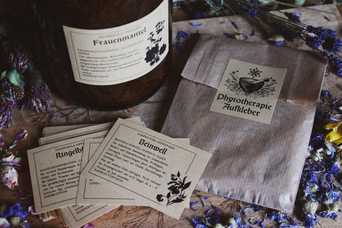 Das Bild zeigt eine Sammlung von Etiketten und Verpackungen für Heilkräuter. Im Mittelpunkt steht eine große braune Flasche mit der Aufschrift „Frauenmantel.“ Außerdem gibt es eine braune Papiertüte mit dem Etikett „Phytotherapie Aufkleber“ und mehrere kleine Karten mit Informationen zu Kräutern wie „Ringelblume“ (Calendula officinalis) und „Beinwell“ (Symphytum officinale).
Die Szene ist auf einer Holzoberfläche arrangiert, umgeben von verstreuten getrockneten Blumen.