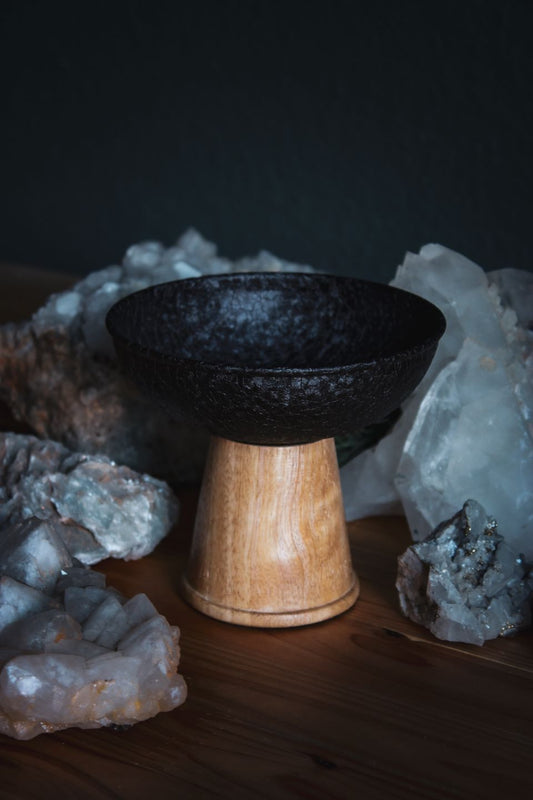 Auf einem Holztisch steht ein schlicht-eleganter und moderner Räucher-Kelch. Sein Körper ist aus schwarzem pulverbeschichteten Eisen. Und sein Fuß ist aus Mangoholz. Um ihn herum stehen verschiedene weiße Mineralien und Steine.