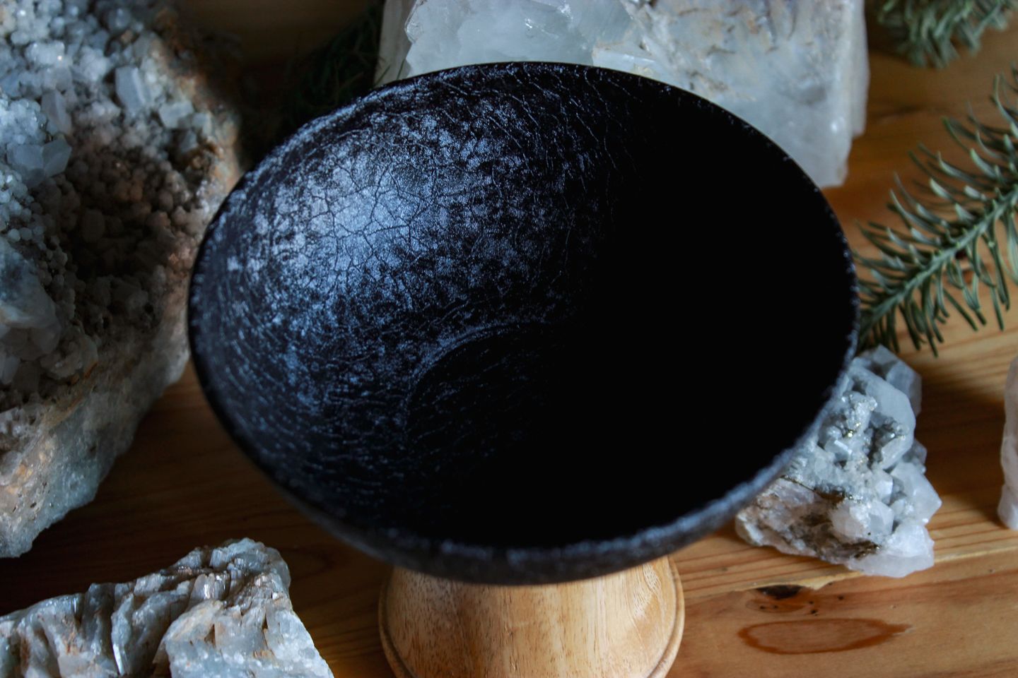 Nahaufnahme des Kelchs von oben: Auf einem Holztisch steht ein schlicht-eleganter und moderner Räucher-Kelch. Sein Körper ist aus schwarzem pulverbeschichteten Eisen. Und sein Fuß ist aus Mangoholz. Um ihn herum stehen verschiedene weiße Mineralien und Steine.